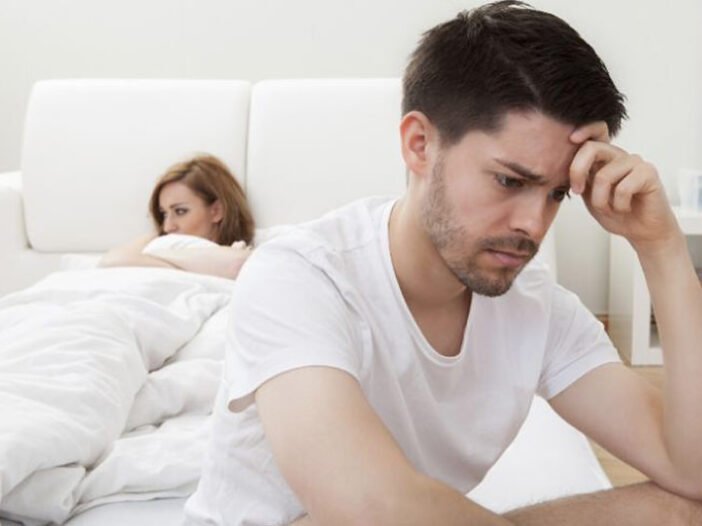 Homem preocupado sentado na beira da cama com sua jovem esposa dormindo ao fundo